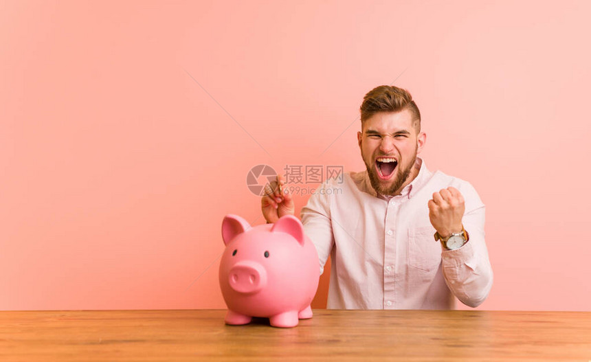 年轻的caucasian男子坐在小猪银行里欢呼着无忧虑和兴奋地欢呼图片