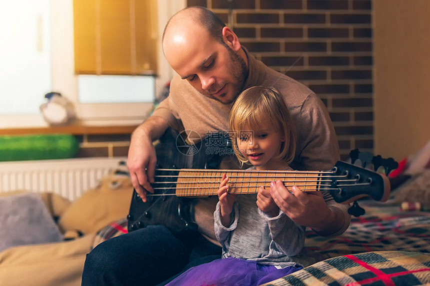 可爱的小女孩和她的父亲坐在家里的沙发上弹着低音吉他和微笑花时间在一起图片