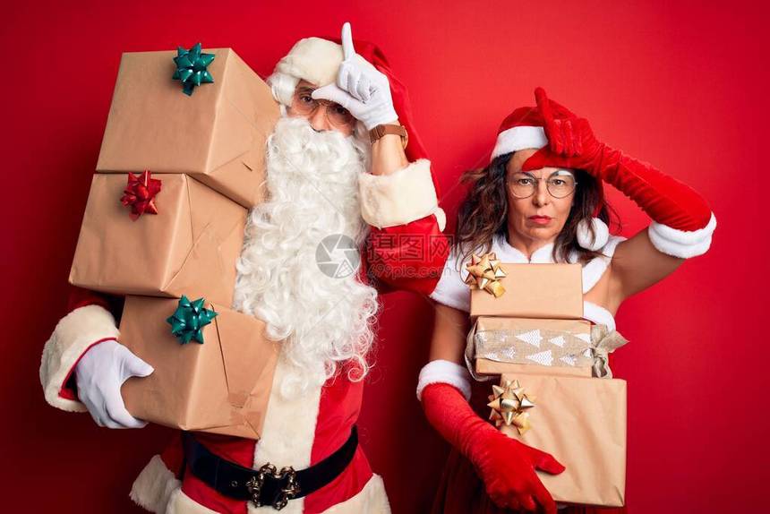 身着圣诞老人服装的中年夫妇持有超乎孤立的红色背景的礼物塔图片