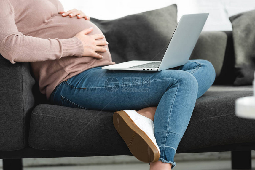 怀孕妇女触摸肚子和在家中用笔记本电脑与医生进行在线咨询的切图片