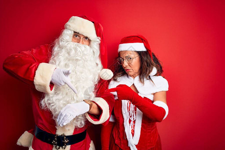 中年夫妇穿着圣诞老人服装和眼镜在孤立的红色背景中匆忙指着看时间图片