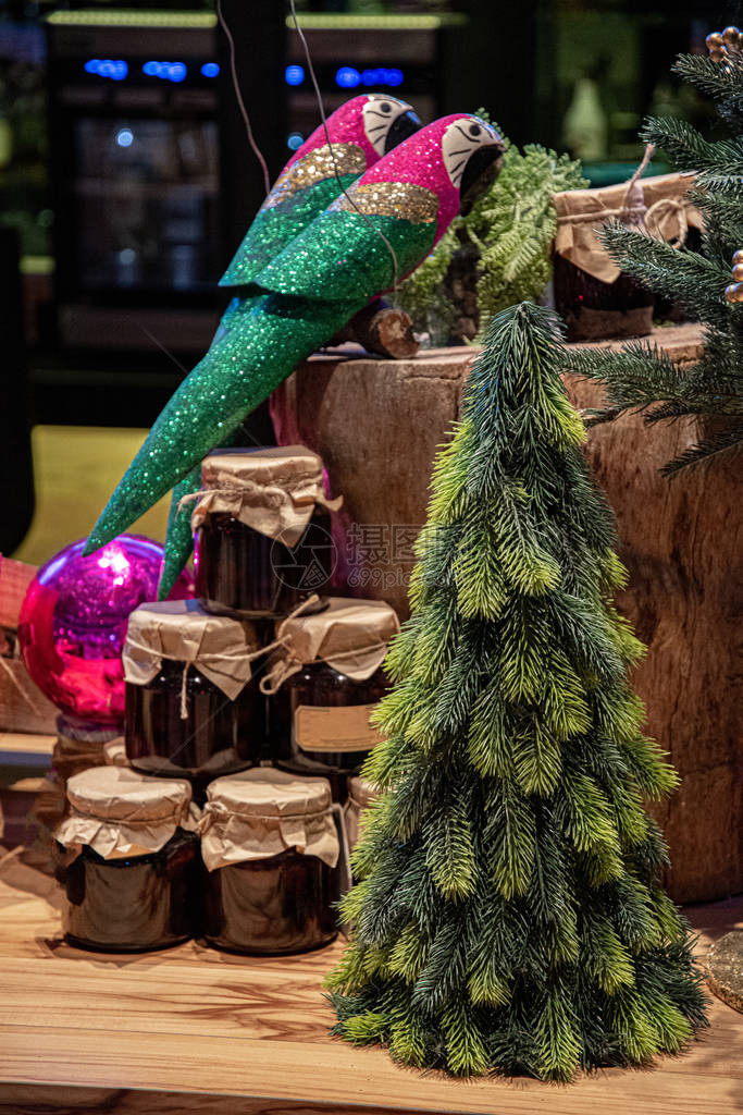 圣诞节装饰木树桩上的两只闪发光的玩具鹦鹉和装有自制果酱的玻璃罐金字塔在前景的人为绿色圣诞树冬季节日装饰品图片