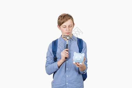 沮丧的学生男孩怀疑打破他的存钱罐使用孤立在白色背景的锤子图片