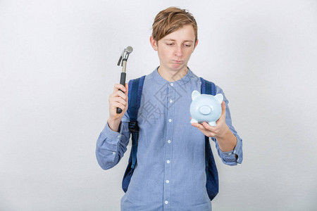 沮丧的学生男孩试图用在白色背景上孤立的锤子打破存钱罐图片