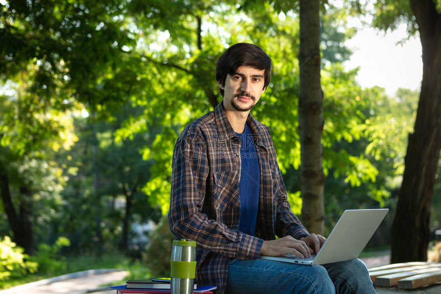 长着胡子和胡子的棕发青年自由职业者与笔记本电脑坐在公园里图片