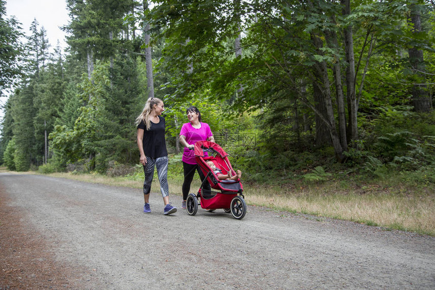 两名30多岁的妇女推着婴儿车在小路上一起散步和交谈沿着美丽的风景秀丽的图片