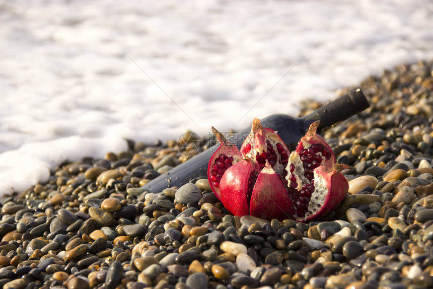 成熟的石榴果与海滩上的瓶酒在海边浪漫的一天背景是海浪和大海水泡沫婚礼或情人节庆图片
