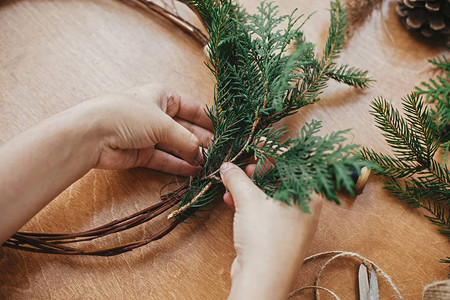 手拿着雪松树枝和松果线剪刀在木桌上制作圣诞花环车间的详细信息制作质图片