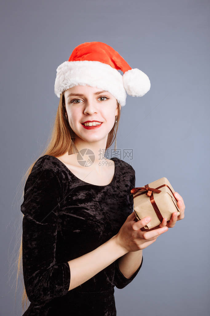 穿红色圣诞帽和黑色礼服的年轻天主教女以灰色背景送给你一个礼物图片