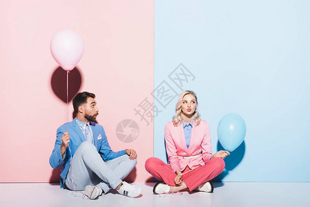 迷人的女人和震惊的男子拿着粉红和蓝底气球在粉色背景