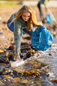 年轻女子从岩石岸边捡垃圾志愿者在阳光明媚的日子清洁海滩她正在保护大图片
