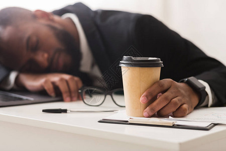 喝咖啡和在工作场所睡觉的精疲力尽的非洲商业办事员图片
