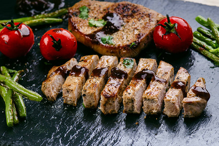 烤猪排牛排配蔬菜西红柿豆类和黑石板上的酱汁带泡沫的鲜肉深色背景烤图片