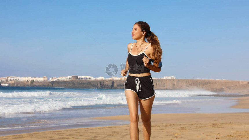 正在夏日海滩上用触摸屏和耳机收听手机臂带音乐的女跑步妇女图片