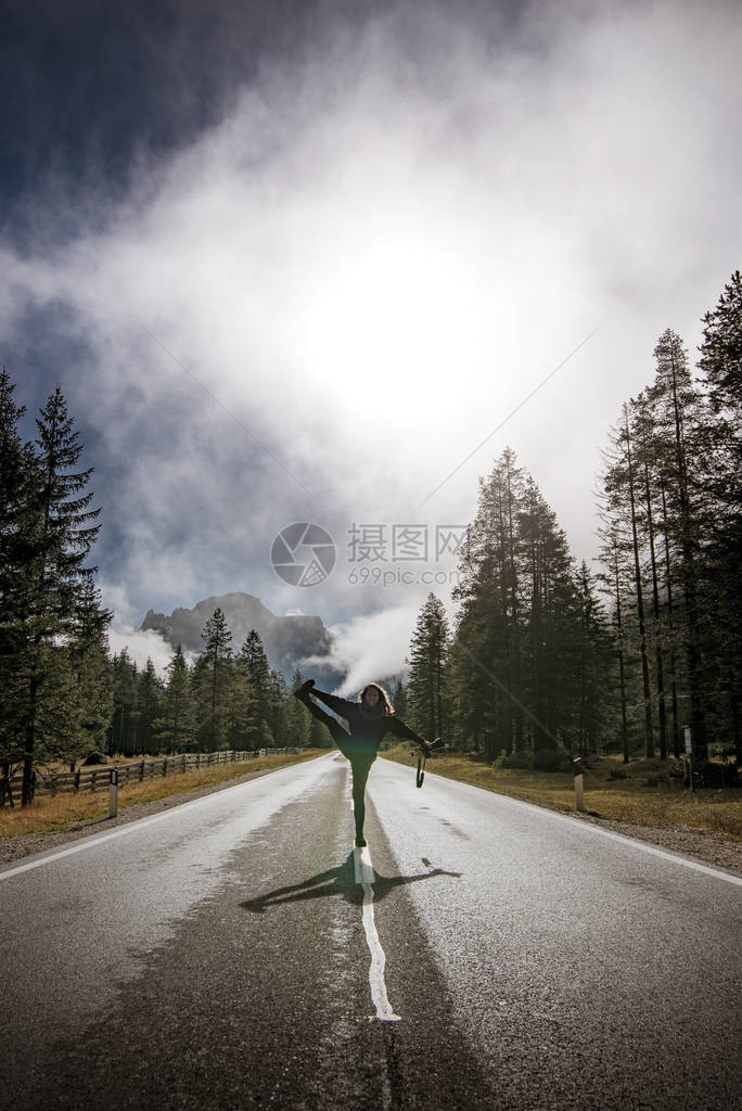 身着黑色夹克在意大利阿尔卑斯山多洛米特山脉沥青路上摆布的年轻女摄影师图片