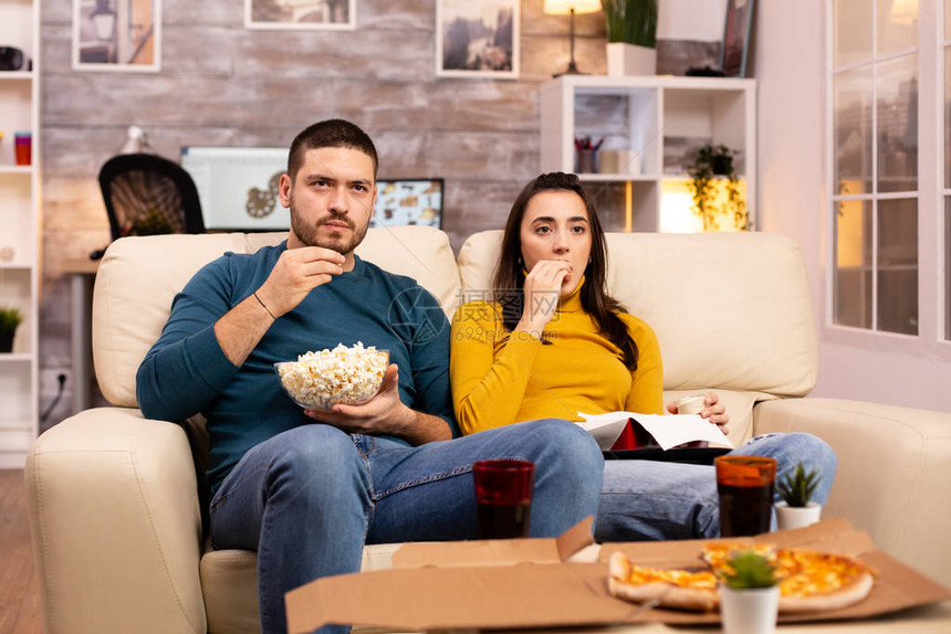 美丽的年轻夫妇在客厅看电视和吃快图片