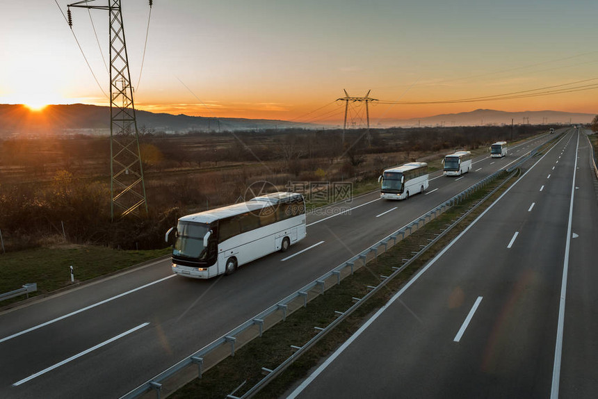 大篷车或四辆白色巴士的车队在令人惊叹的橙色日落天空下的乡村公路上行驶与白色公共汽图片