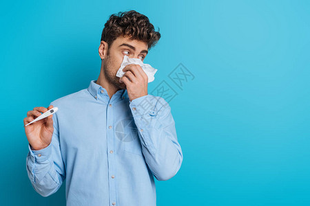 患病的年轻人用纸巾擦鼻子在蓝色背景背景图片