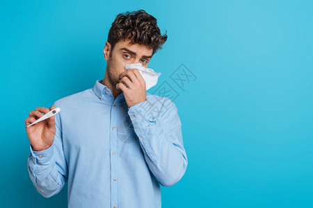 年轻病人用纸巾擦鼻涕和在蓝底显示温度图片