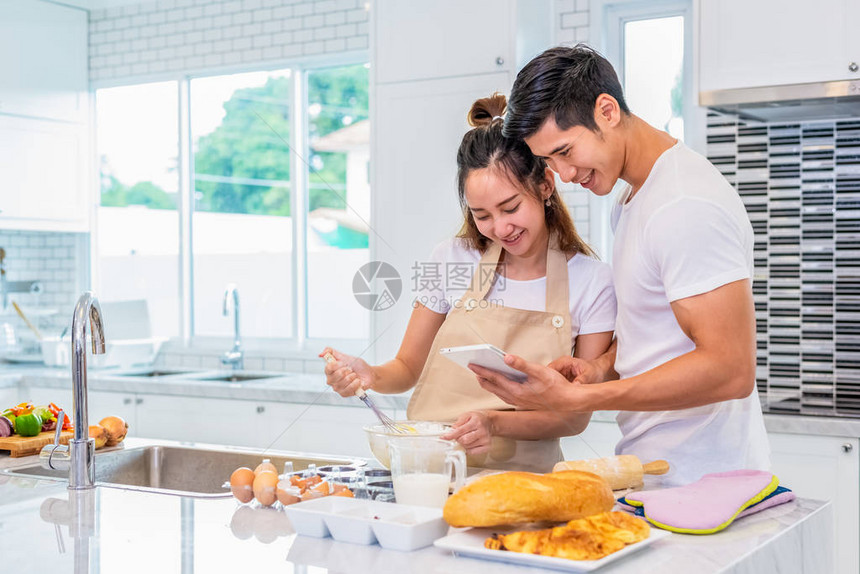 快乐的亚洲夫妇在厨房里一起做饭和烤蛋糕寻找平板电脑的男人和女人在家里按照食谱步骤爱与幸福的概念甜蜜月图片