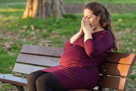 妊娠剧吐年轻的孕妇在温暖的阳光下坐在公园的长椅上背景