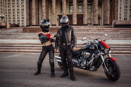 美丽的夫妇在莫斯科一座美丽的庄严建筑的背景下骑着一辆很酷的摩托车穿着摩托车服的野蛮而严肃的家伙和一个温柔时尚的女背景图片