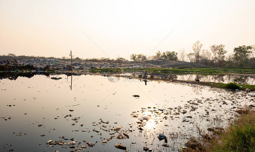 污染的水和山大垃圾堆和太阳的污染在背景中落下图片