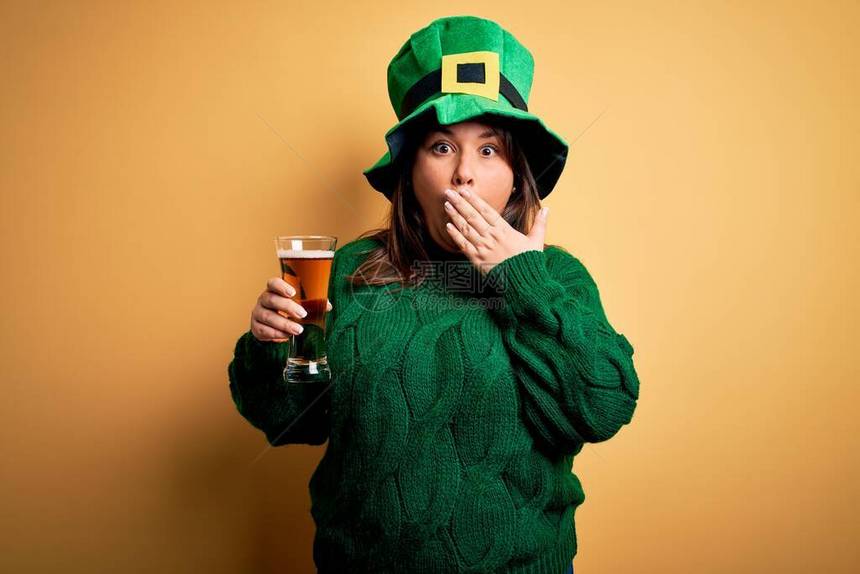 年轻漂亮的大码女人戴着绿色帽子庆祝圣帕特里克节喝啤酒盖嘴用手震惊为错误感到羞耻图片