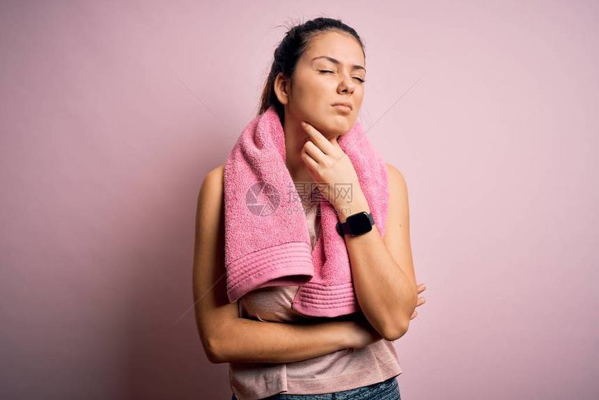 年轻漂亮的褐发女运动员穿着运动服和粉红色背景的毛巾触摸颈部疼痛喉咙痛以致流感图片