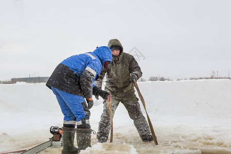 穿着冬装头戴兜帽的工人在结冰的河冰上的冰块下制作吊索图片