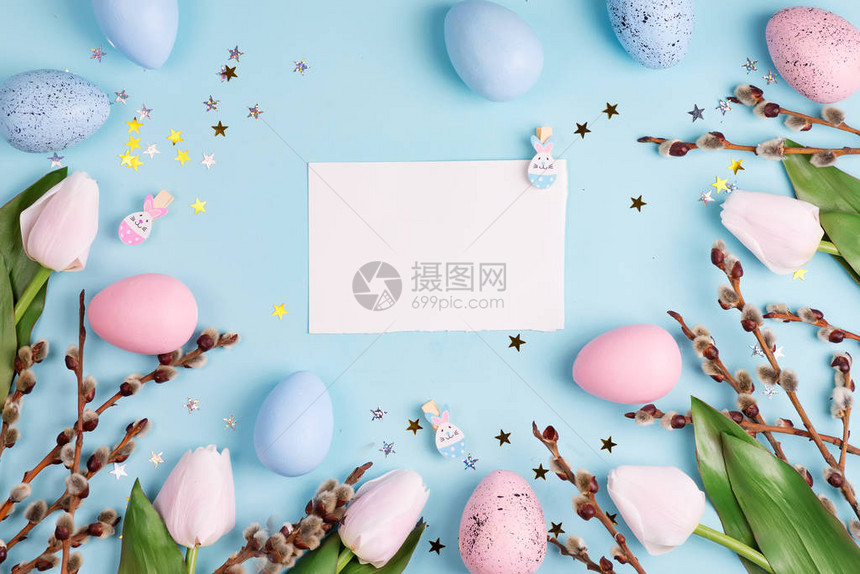 复活节柳花环白色郁金香和蓝色背景上的蓝色粉红色复活节彩蛋与纸顶视图片