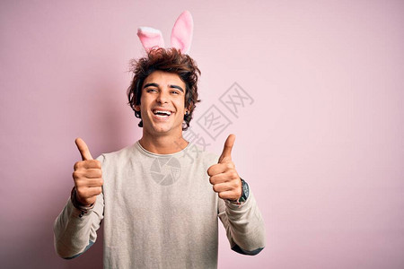 年轻英俊的男人拿着复活节兔子耳朵站在孤立的粉红色背景成功标志上图片