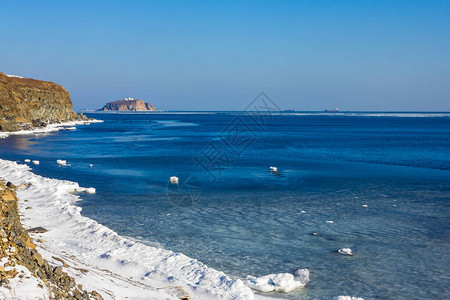 岩石海岸被白雪覆盖平静的水面明亮的蓝色远处是一座石岛晴朗阳图片