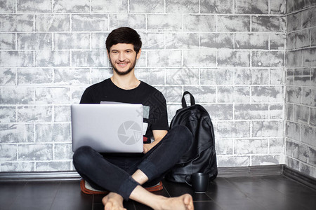 坐在地板上的年轻人手拿着笔记本电脑和黑杯咖啡或茶灰砖墙的背景图片