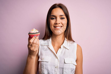 年轻漂亮的黑发女人在孤立的粉红色背景中吃巧克力纸杯蛋糕图片