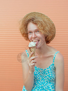 年轻开朗的卷发红女人在草帽和蓝色夏装吃米色背景上的冰淇淋图片