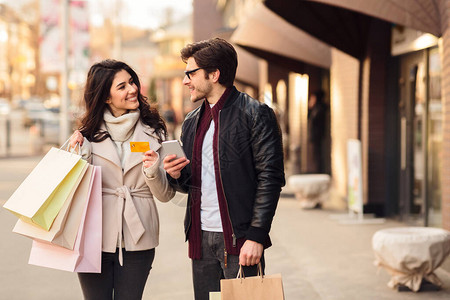 可爱的夫妇在市中心露天户外行走时拥有智能手机和信用卡图片