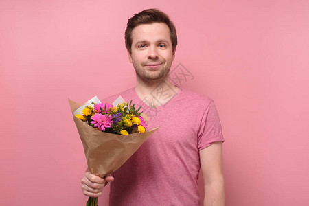 身穿粉色t恤的年轻白人男子拿着鲜花作为生日礼物送给母亲或女朋友周年纪念礼物的概念在彩色墙上背景图片
