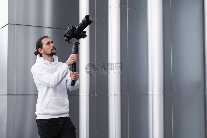 年轻的专业摄像师在3轴云台稳定器上手持专业相机专业设备有助于制作高质量视频而不会晃动摄影师穿着白色连图片