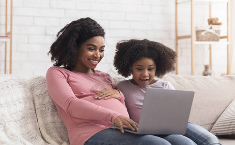 家庭休闲年轻怀孕的非洲女青年以及她可爱的小女儿在笔记本电脑上看电影图片