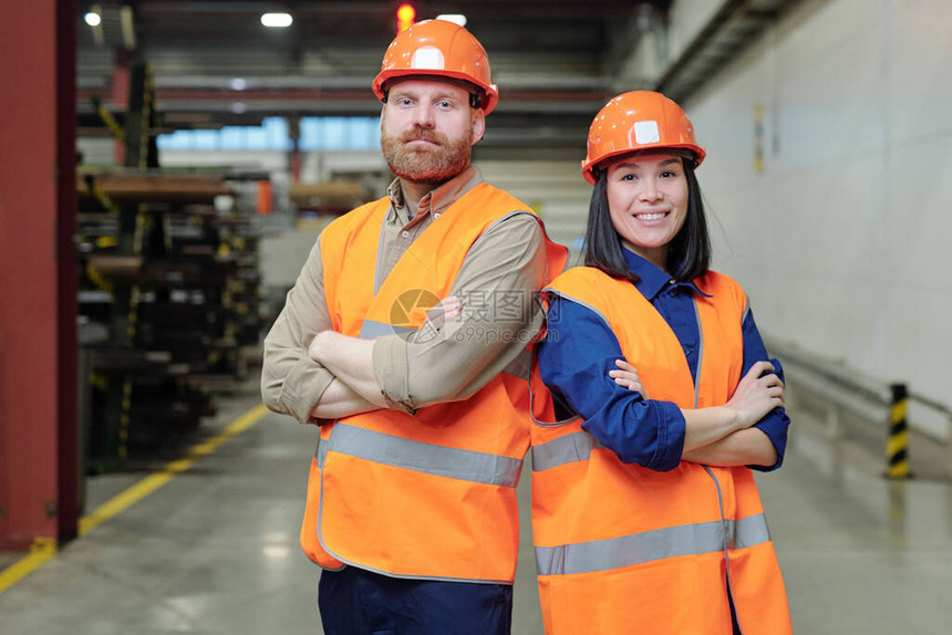 两名在硬哈特和工作服中成功的年轻工程师站在工厂里时胸交叉手臂图片
