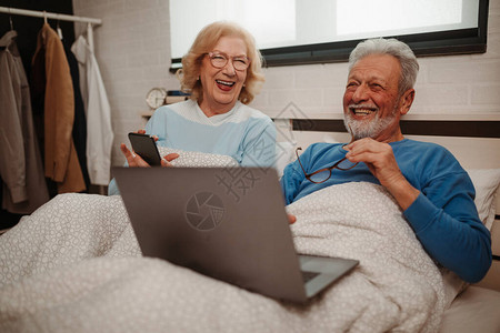 老年夫妇躺在床上看着他们的智能手机的照片老人拿着他的笔记本电脑正图片