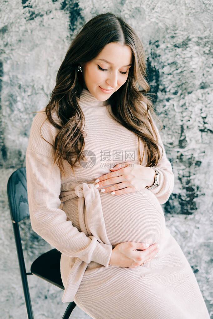 一位身着米色连衣裙的年轻漂亮孕妇坐在靠近灰色墙壁的椅子图片