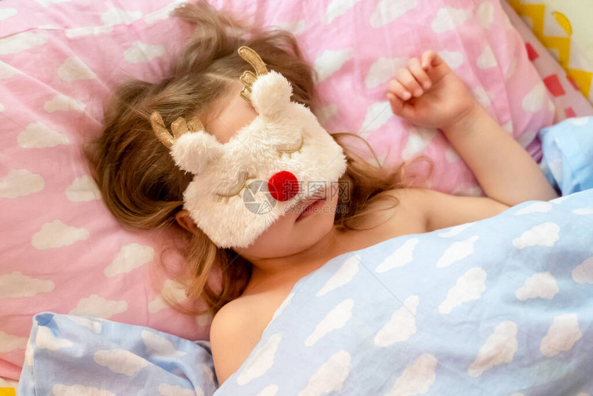 小女孩戴着有趣的鹿形毛绒面具安静地睡在床上就寝时间孩子睡在有云的床单上图片