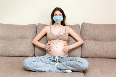 身戴保护医疗面具的孕妇坐在家里沙发上图片