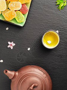 茶壶和装有绿茶的小杯子位于黑色石桌上背景上的樱花和瓣糖果图片