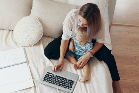 年轻母亲及其孩子在家卧室用笔记本电脑做家务图片