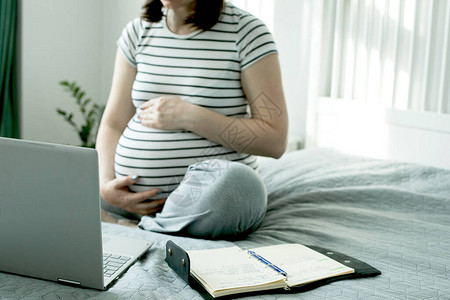 美丽怀孕的年轻女子坐在卧室的床上用笔记本电脑工作图片