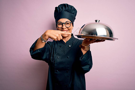 中年黑发女厨师穿着炊具制服和帽子拿着托盘圆顶非常高兴地用图片