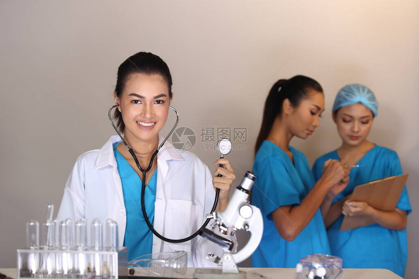 穿白衬衫听诊镜的美丽的亚洲女医生展示了注射器和两个护士病历表Hygine帽子模糊不清图片